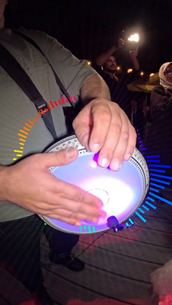 Person playing a round hand drum with a glowing purple light, at night, as others watch and a photographer captures the moment.